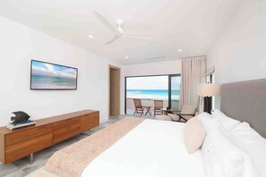 Une chambre d’hôtel moderne avec un grand lit, une commode en bois, un ventilateur de plafond et une vue sur la plage, visible à travers une grande fenêtre.
