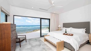 A modern bedroom with a large window overlooking the ocean, a bed with a tray of wine, and a ceiling fan. - Halcyon Point: New Luxurious Oceanfront Villa w/ Pool, Spa + Tanning Terrace (Grand Cayman)