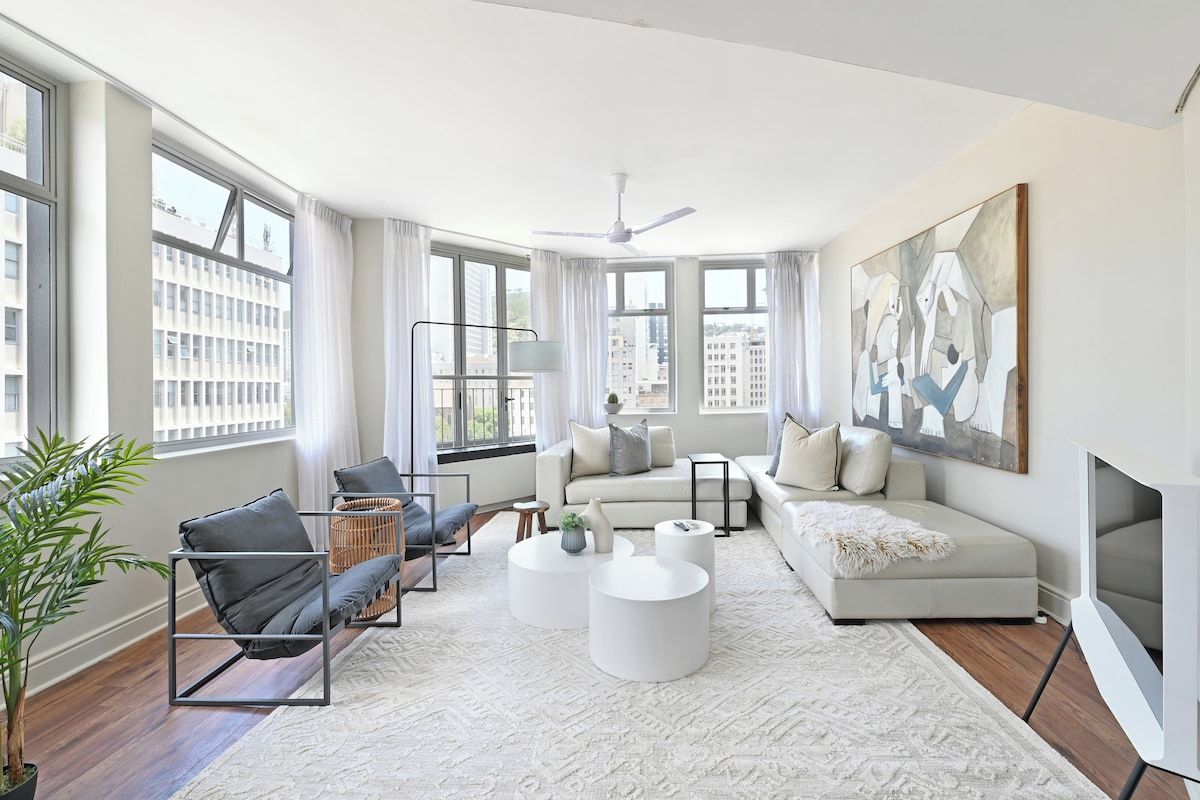 A modern living room with a sofa, armchairs, a coffee table, and a large window offering a city view.
