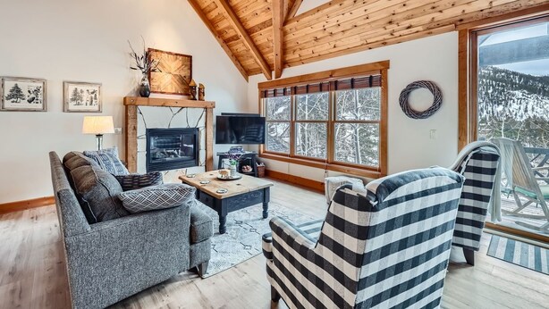 Ein gemütliches Wohnzimmer mit Kamin, einer Couch, gestreiften Sesseln und einem großen Fenster mit Blick auf schneebedeckte Bäume.