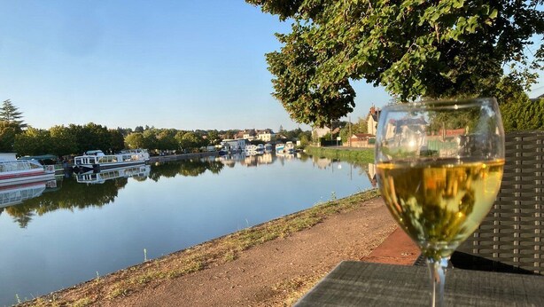 Ein Glas Weißwein auf einem Tisch mit Blick auf einen ruhigen Fluss und Boote im Hintergrund.