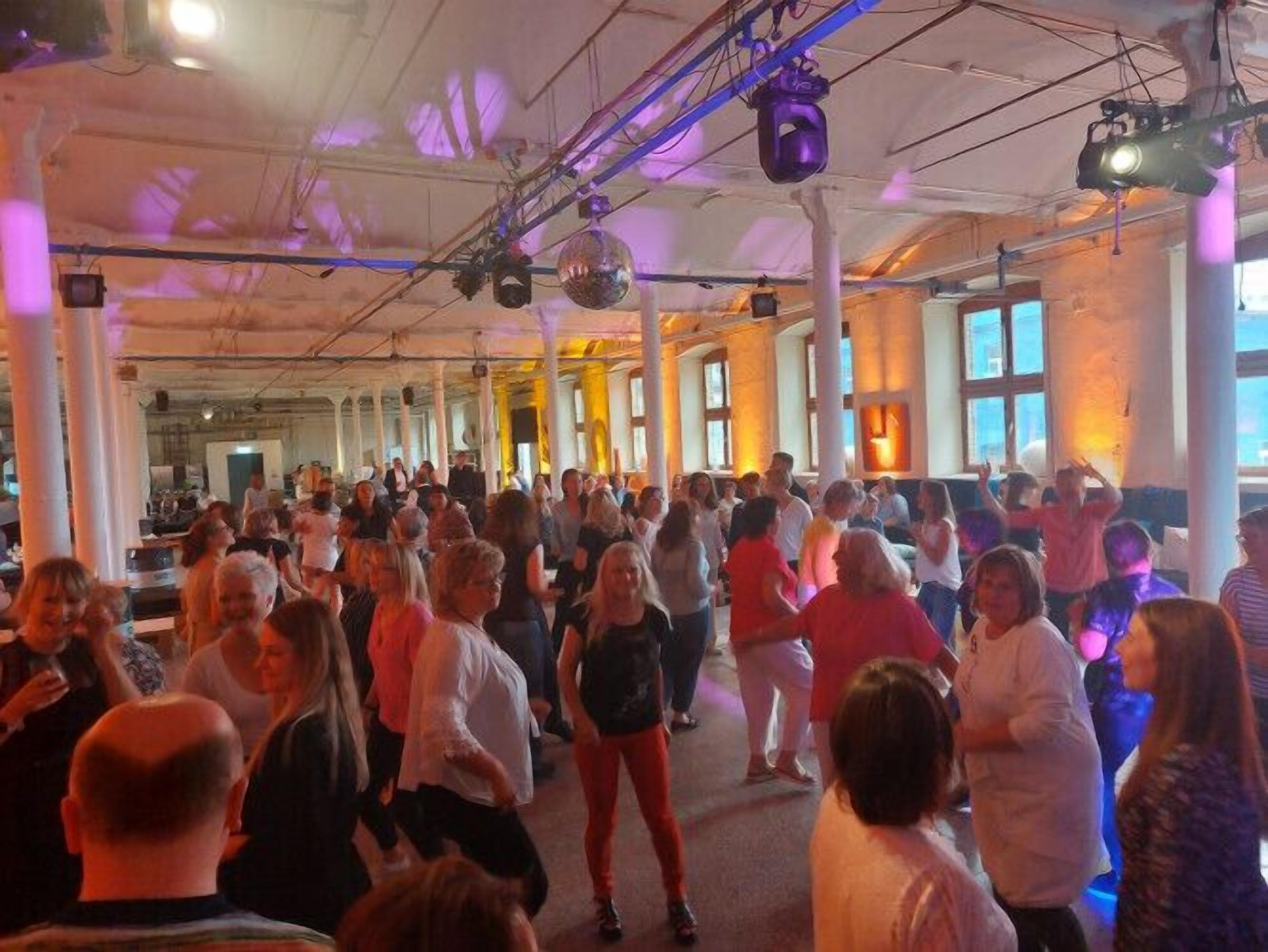 A spacious hall with high ceilings and large windows, filled with people dancing.