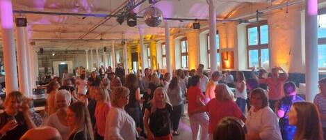 A spacious hall with high ceilings and large windows, filled with people dancing.