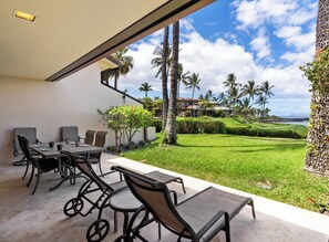 Een terras buiten met een tafel en stoelen, uitzicht op een verzorgd gazon, palmbomen en een verre huizen en oceaan.