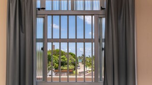 A window with metal bars and grey curtains, offering a view of a residential area with trees and parked cars. - AP 37 Ala Canto Gran Residencial Solavir (Bombinhas)