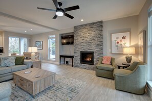 A spacious living room with a stone fireplace, a ceiling fan, and a large TV. - Gardener's Getaway - Oyhut Bay Seaside Resort (Ocean Shores)