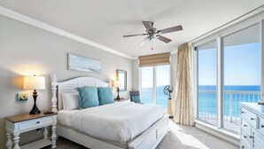 A bedroom with a bed, bedside tables, a ceiling fan, and a large window with a view of the ocean.