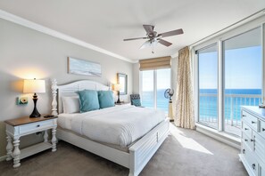 Une chambre à coucher comprenant un lit, des tables de chevet, un ventilateur de plafond et une grande fenêtre donnant sur l’océan.
