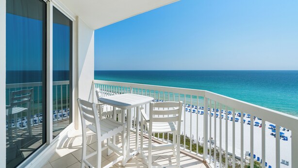A balcony with white chairs and a table, overlooking a beach with blue umbrellas and a clear sky.