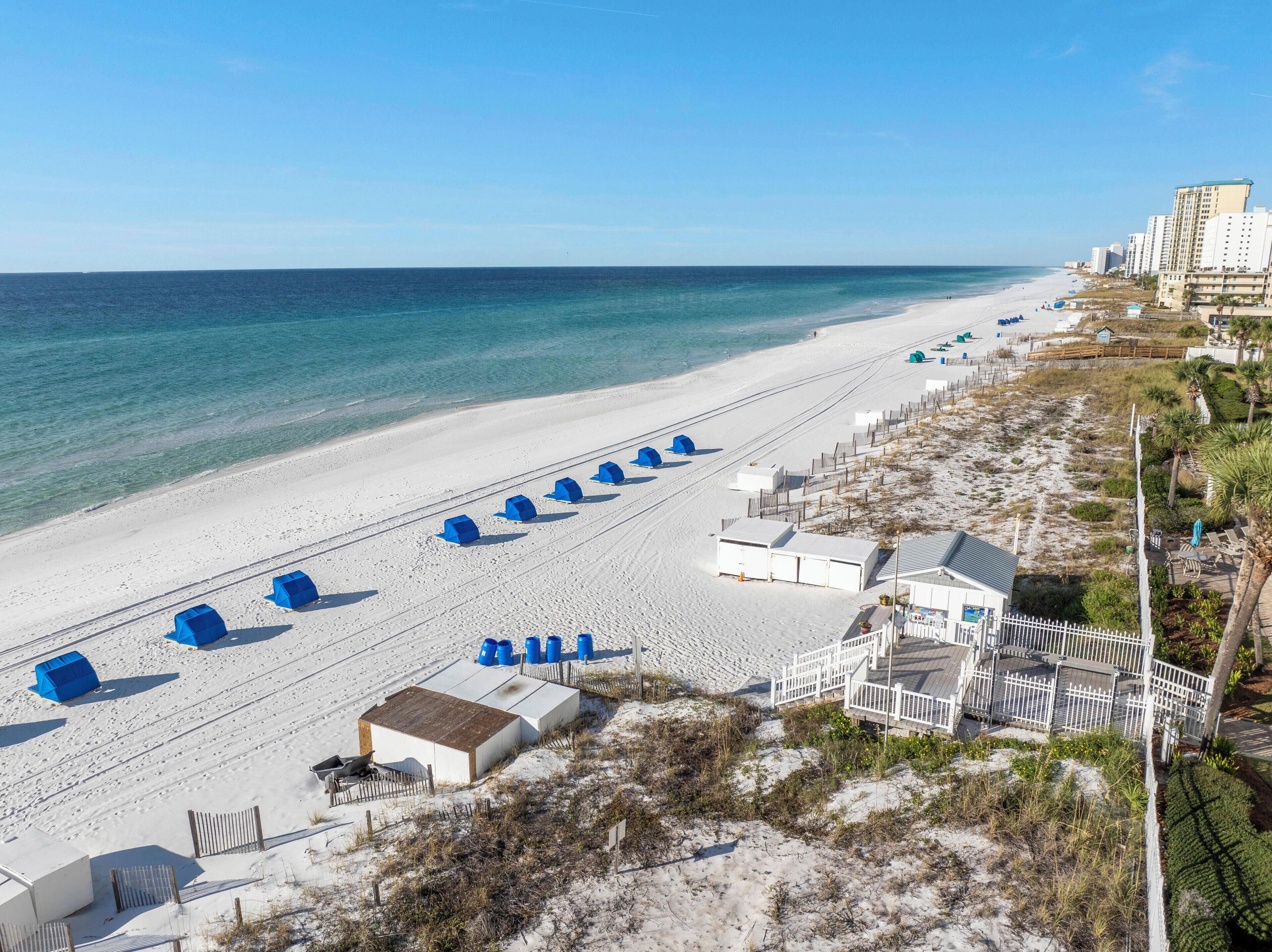 A beach with white sand, a row of blue beach huts, and a white building with a fenced deck.