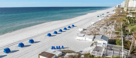 A beach with white sand, a row of blue beach huts, and a white building with a fenced deck.