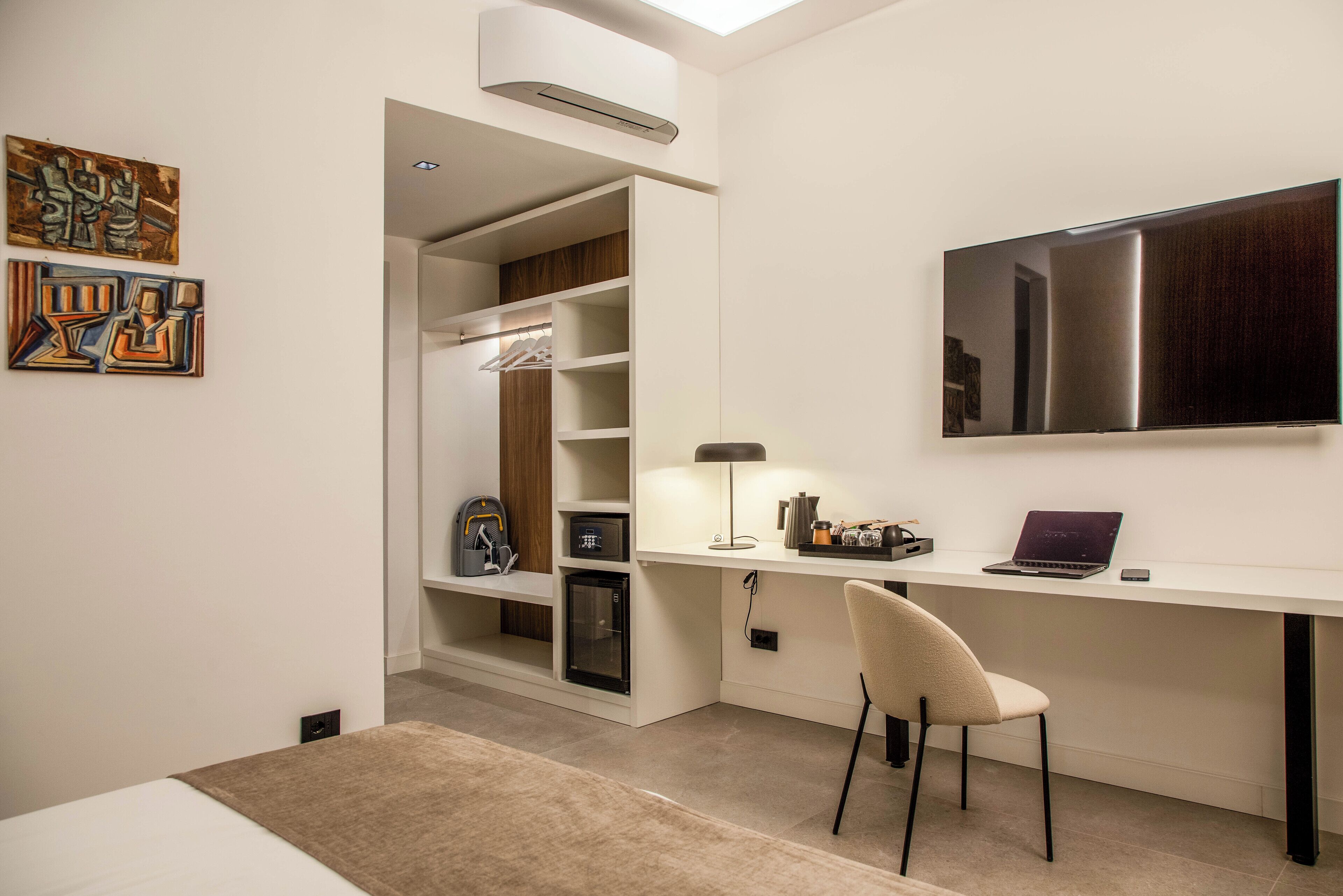 Une chambre à coucher moderne avec un lit, un bureau avec un ordinateur portable, une chaise, une armoire et des œuvres d’art accrochées au mur.