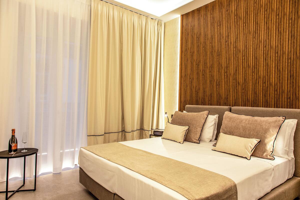 Une chambre d’hôtel avec un grand lit, une literie de couleur beige, un mur décoratif en bois et une petite table sur laquelle se trouvent une bouteille et un verre.