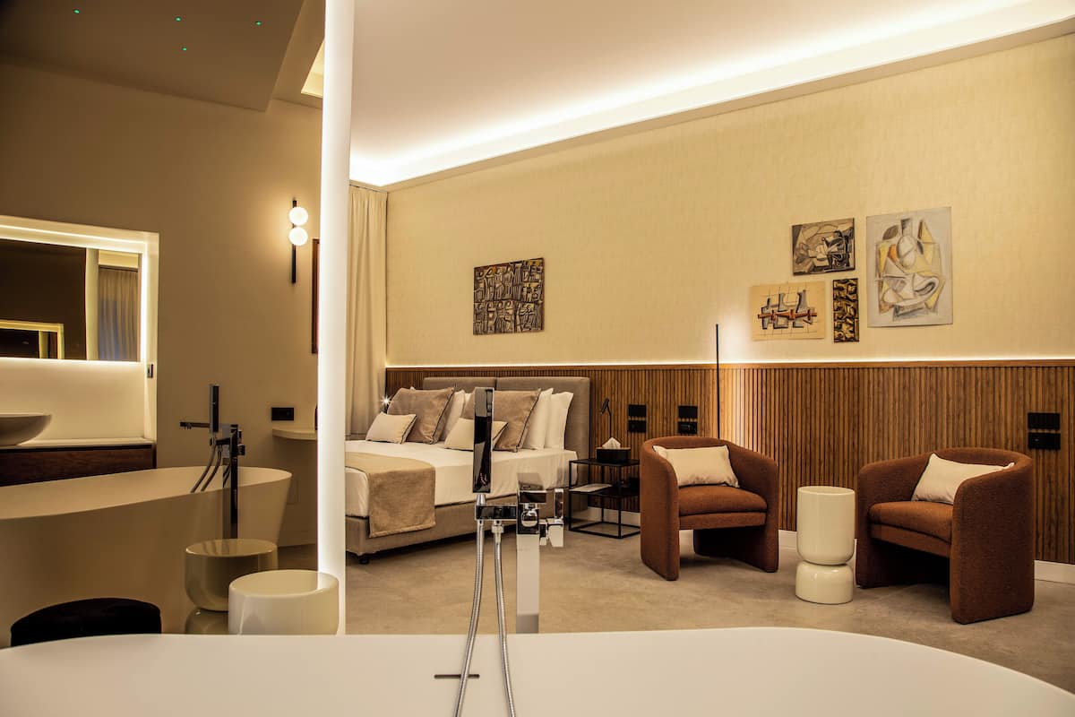 Une chambre d’hôtel moderne dotée d’un grand lit, de deux fauteuils, d’une coiffeuse avec baignoire et d’œuvres d’art accrochées aux murs.
