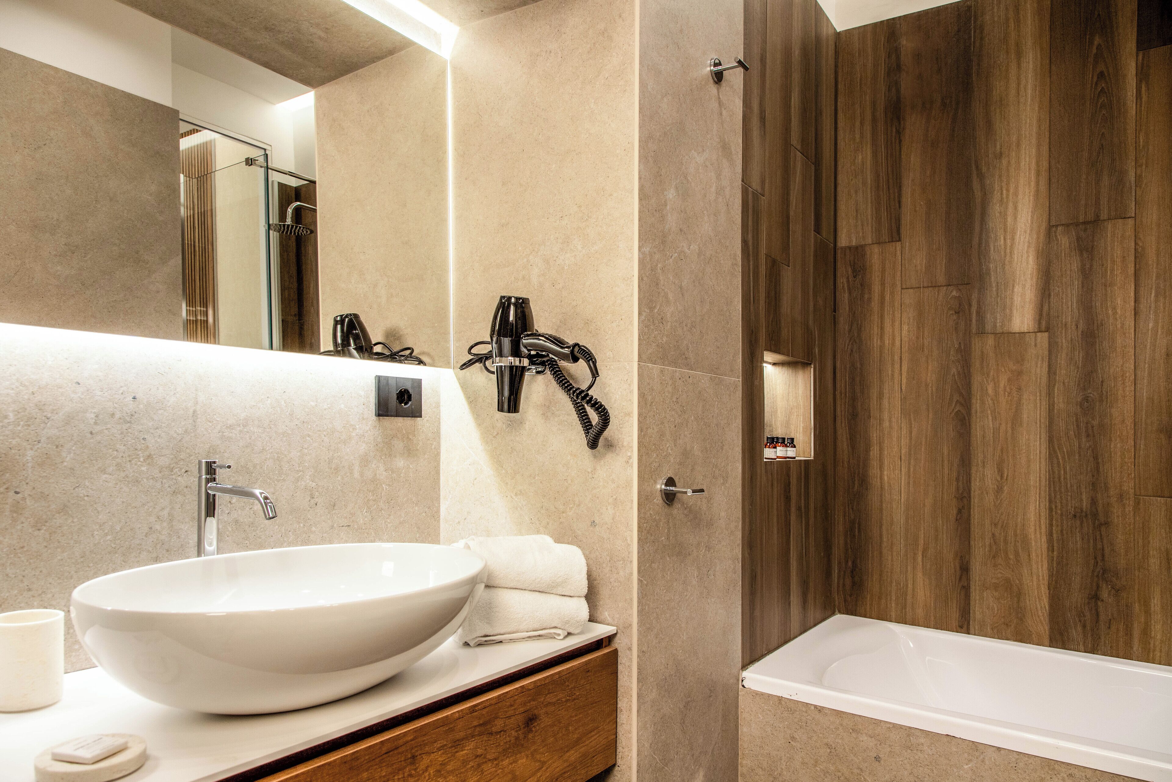 A modern bathroom with a large white vessel sink, a wooden vanity, a glass shower enclosure, and a wooden wall panel.