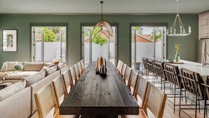 A spacious dining area with a long wooden table and multiple chairs, a cozy seating area with a sofa, and a kitchen with a white refrigerator and a hanging light fixture. - 14BR Luxury Compound | Private Pool, Built-in spa (Savannah)