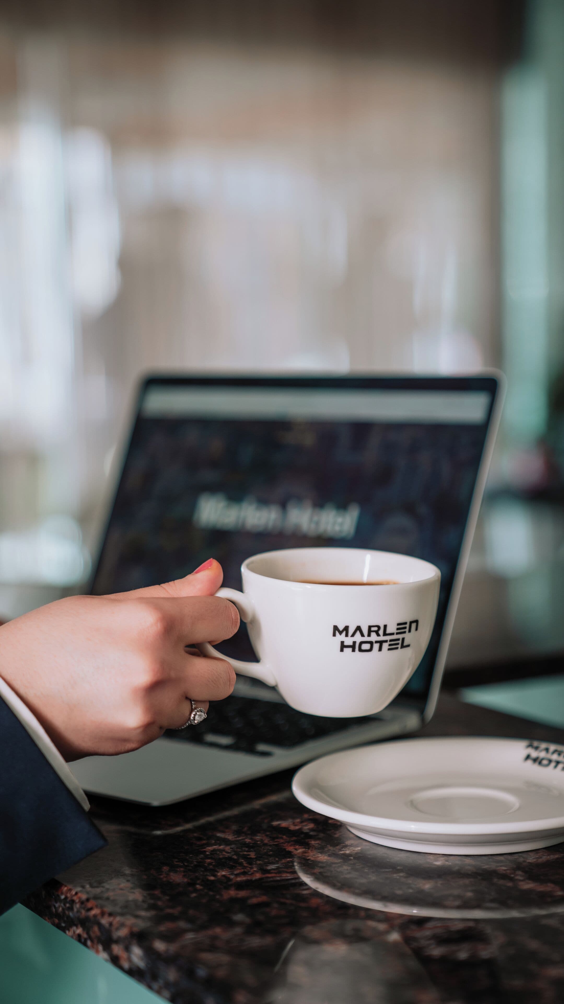 Una mano sosteniendo una taza de café con el texto "MARLEN HOTEL", una laptop mostrando "Marlen Hotel" en la pantalla y un plato sobre la mesa.