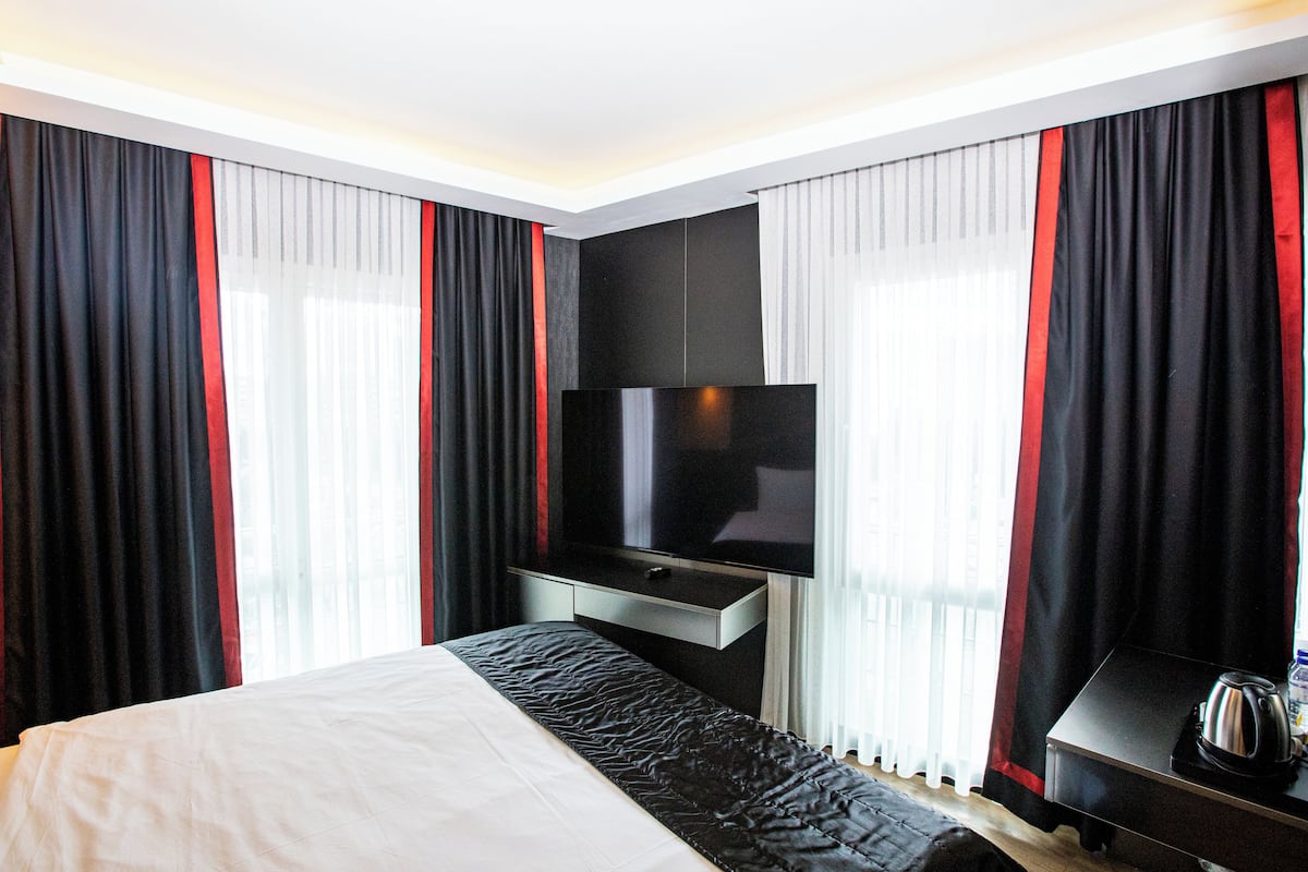 Quarto de hotel com cama, televisão de tela plana na parede, mesa de cabeceira com chaleira e cortinas com detalhes vermelhos.