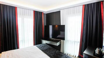 A hotel room with a bed, a flat-screen TV mounted on the wall, a bedside table with a kettle, and curtains with red accents.