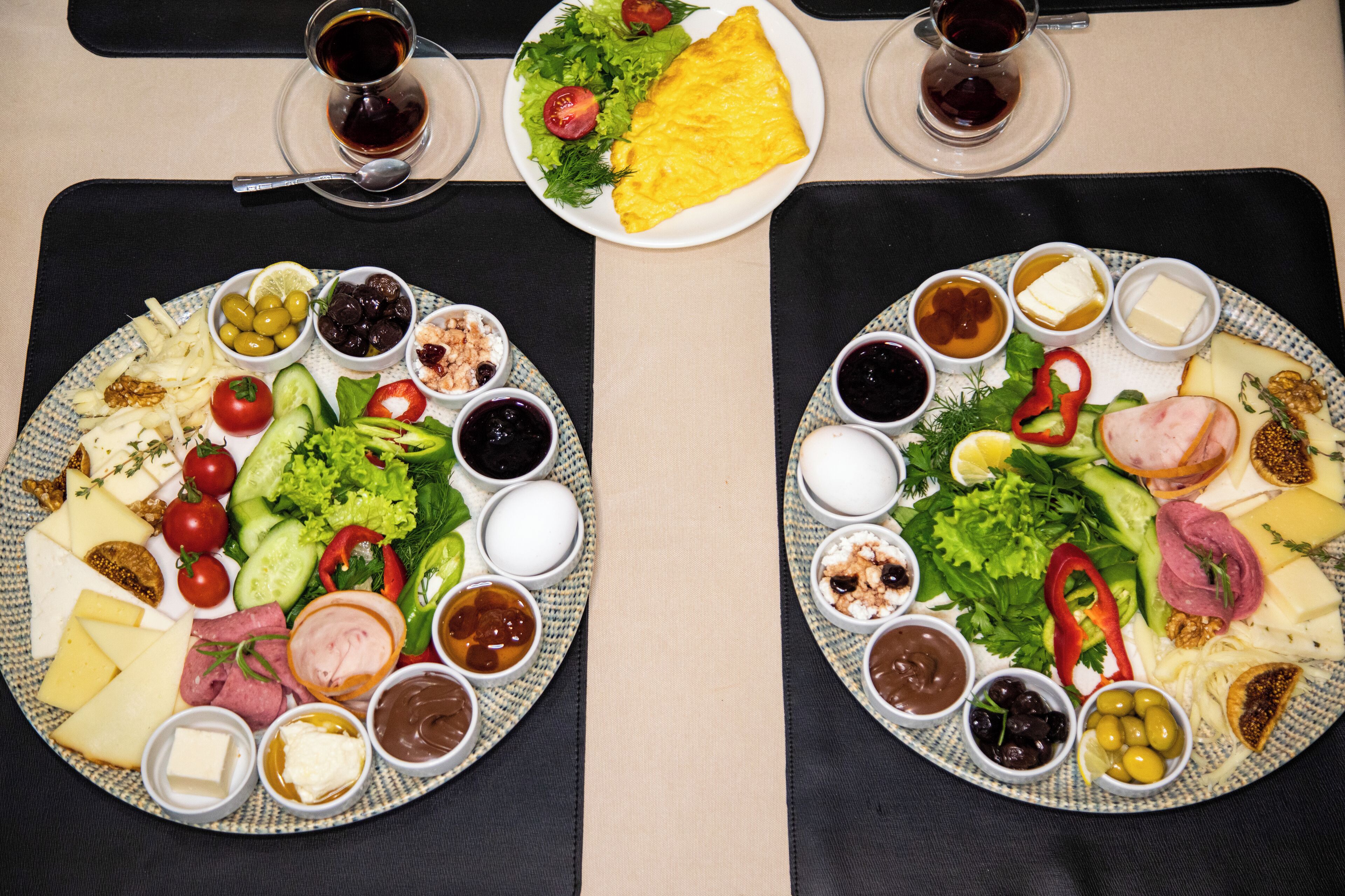 Two plates of food with various cheeses, meats, and vegetables, accompanied by small bowls of condiments and drinks on a table.