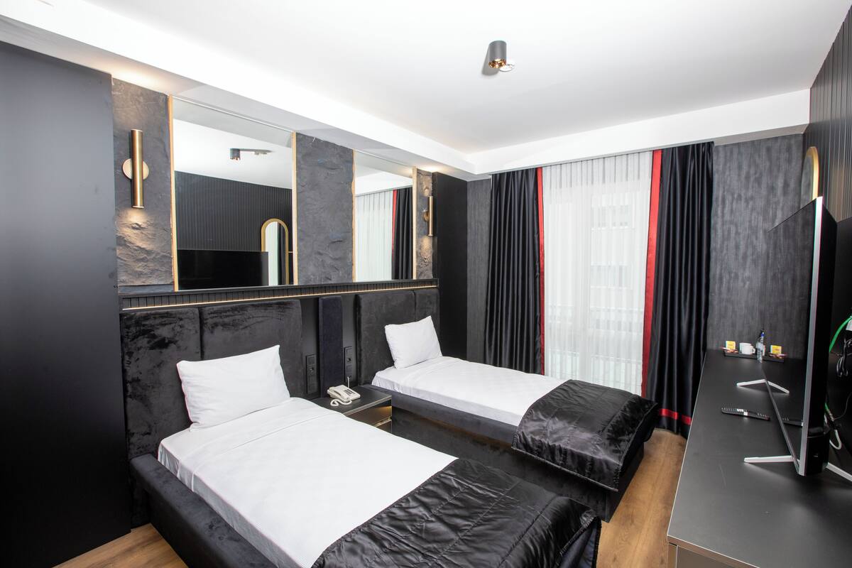 Quarto de hotel moderno com duas camas, um espelho grande, uma escrivaninha com computador e uma janela ampla com cortinas.