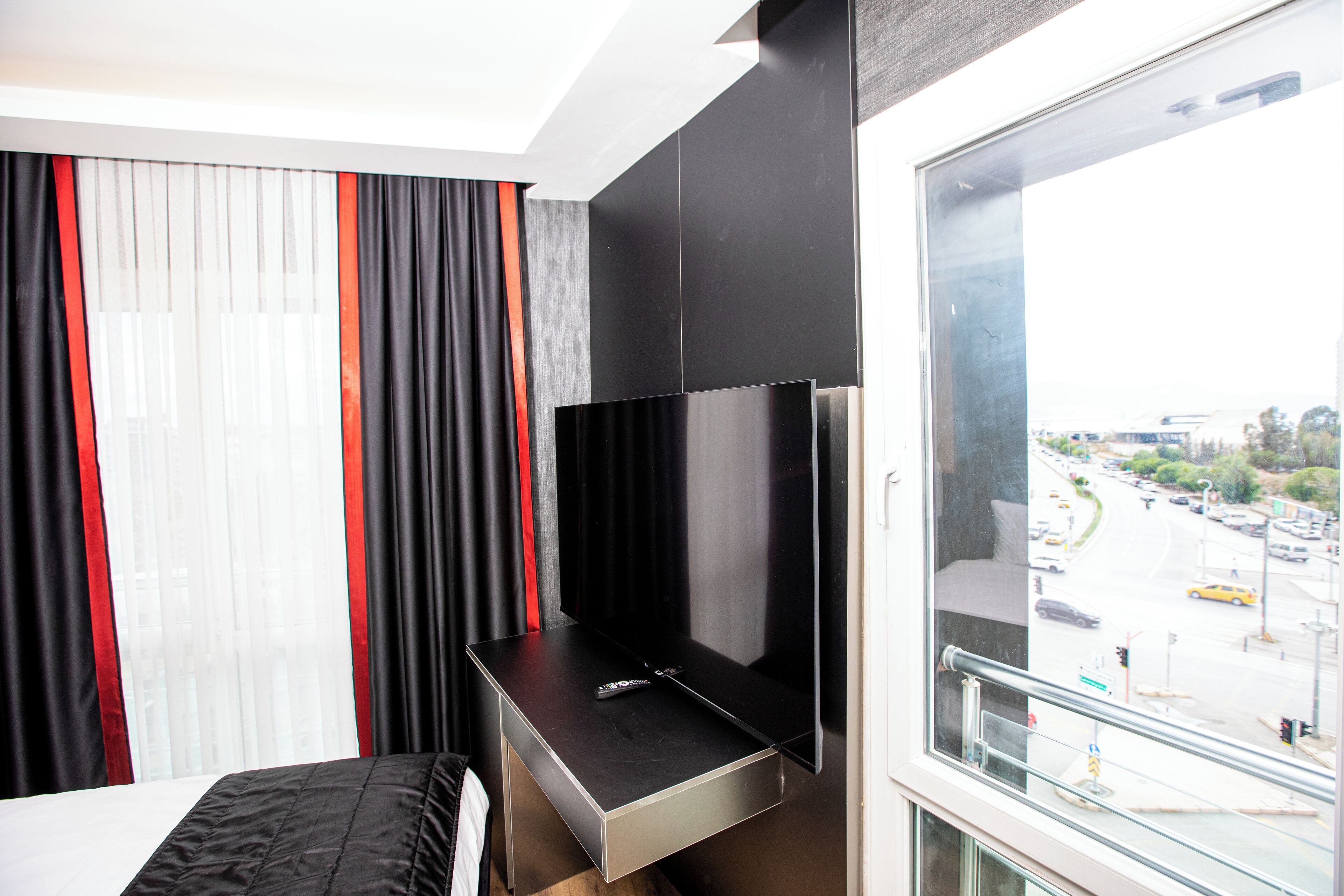 A hotel room with a bed, a TV mounted on a wall, a window with a city view, and a balcony railing.