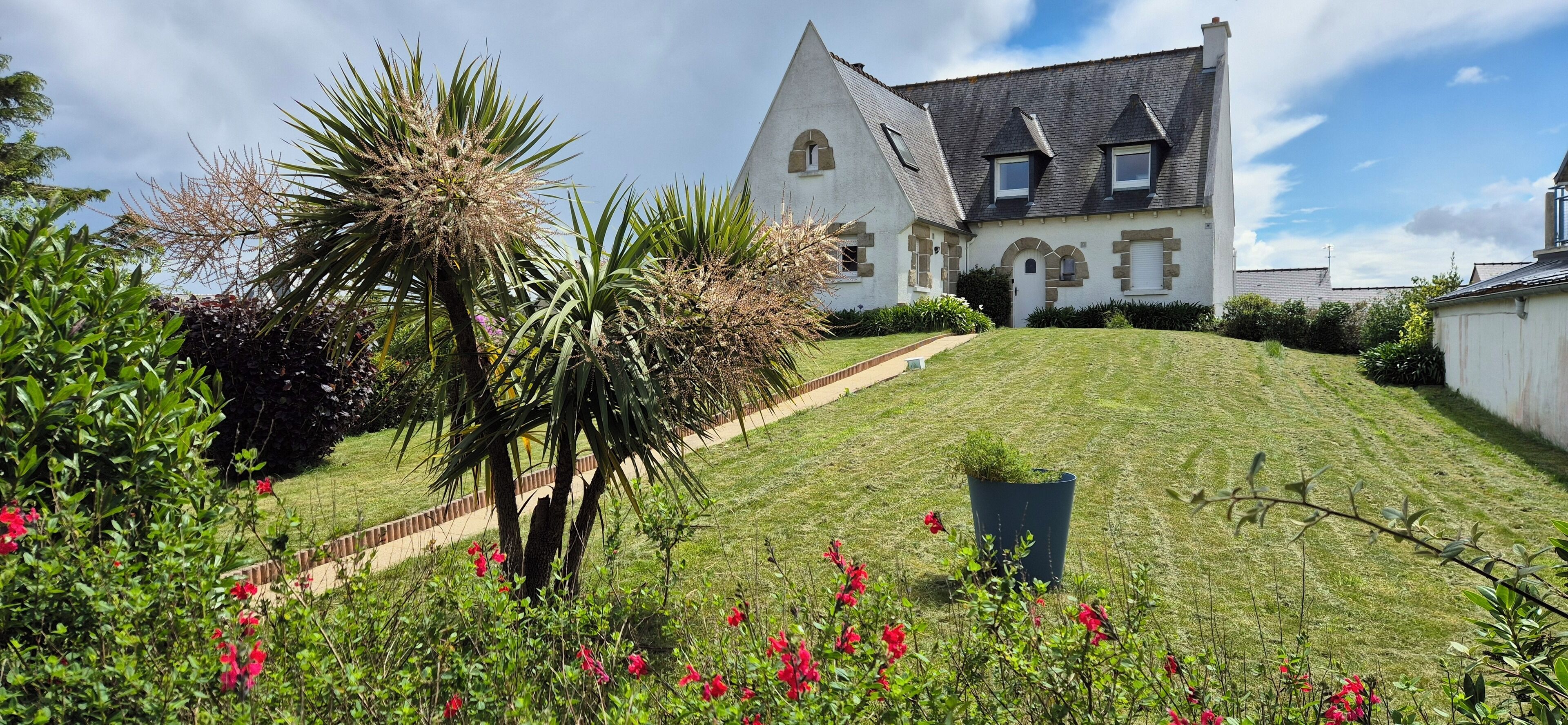 A large, white, two-story house with a steeply pitched roof, surrounded by a well-maintained lawn and flowering plants.