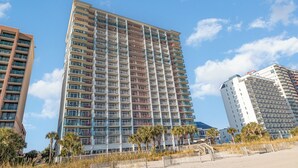 A high-rise hotel with multiple balconies, located near a beach with palm trees and a sandy area. - Spectacular 3 BR 2 BA Ocean Front Condo (Myrtle Beach)