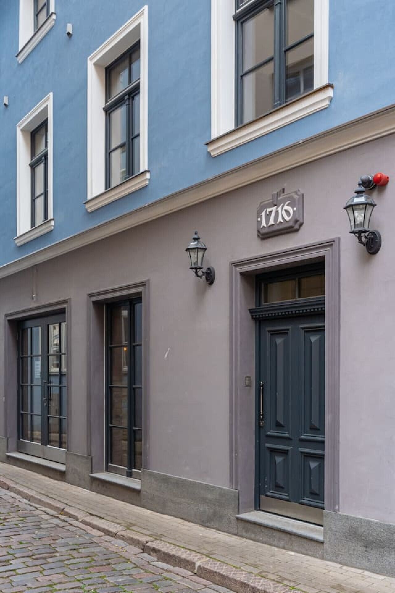 A building with a blue facade, numbered 1716, featuring dark doors and windows with white frames.