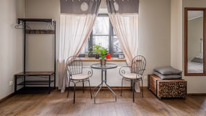 A room with a wooden floor, a small round table with chairs, a radiator, a window with curtains, a hanging shelf, and a mirror on the wall.
