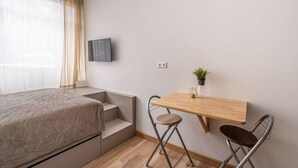 A bedroom with a bed, a TV mounted on the wall, a small wooden desk with a chair, and a potted plant. - Dzintaru studio apartment (Jūrmala)