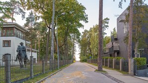 A street lined with trees and a statue, leading to a modern building with balconies.