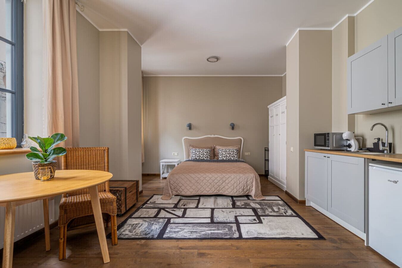 A compact bedroom with a bed, bedside table, chair, and a small kitchenette.