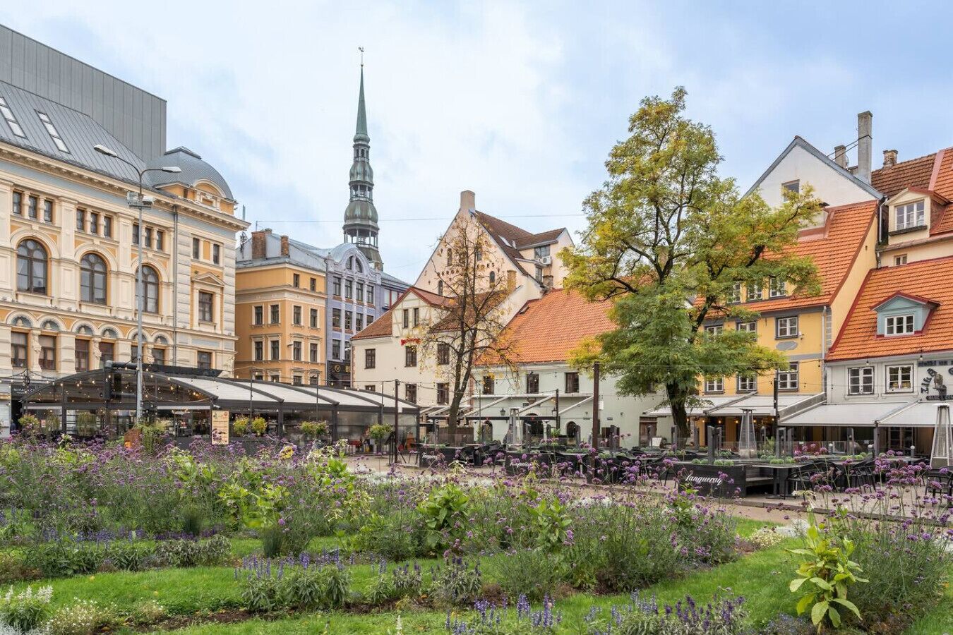 Ett historiskt torg med traditionell arkitektur, ett framträdande kyrktorn och en blomsterträdgård.
