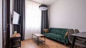 Ein modernes Wohnzimmer mit einem grünen Sofa, einem Holztisch, einem an der Wand befestigten Fernseher und einem Essbereich mit Tisch und Stühlen.