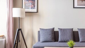 A modern living room with a wooden floor, a grey sofa with cushions, a standing floor lamp, and two framed pictures on the wall.