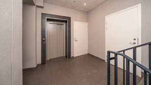 Un passadís amb un ascensor modern, una porta blanca i una escala amb barana metàl·lica.
