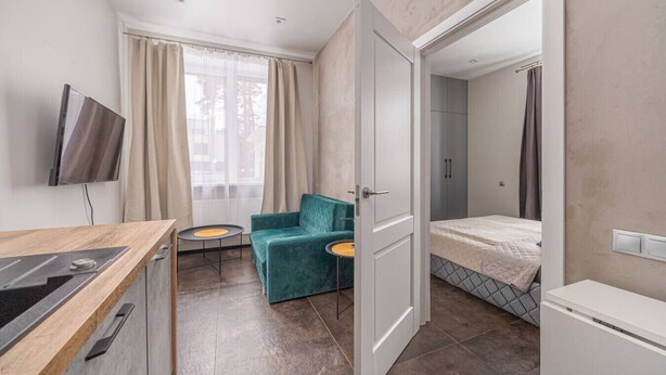 Ein kompaktes Hotelzimmer mit Küchenzeile, einem Einzelbett, einer kleinen Sitzecke mit einem Sessel und einem an der Wand befestigten Fernseher.