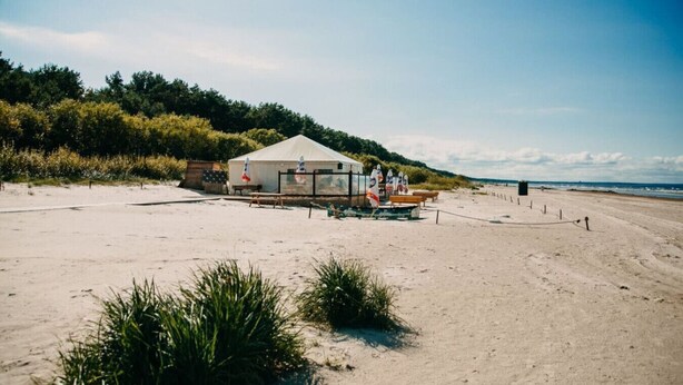 Una estructura junto a la playa, con techo blanco, rodeada de arena y vegetación.