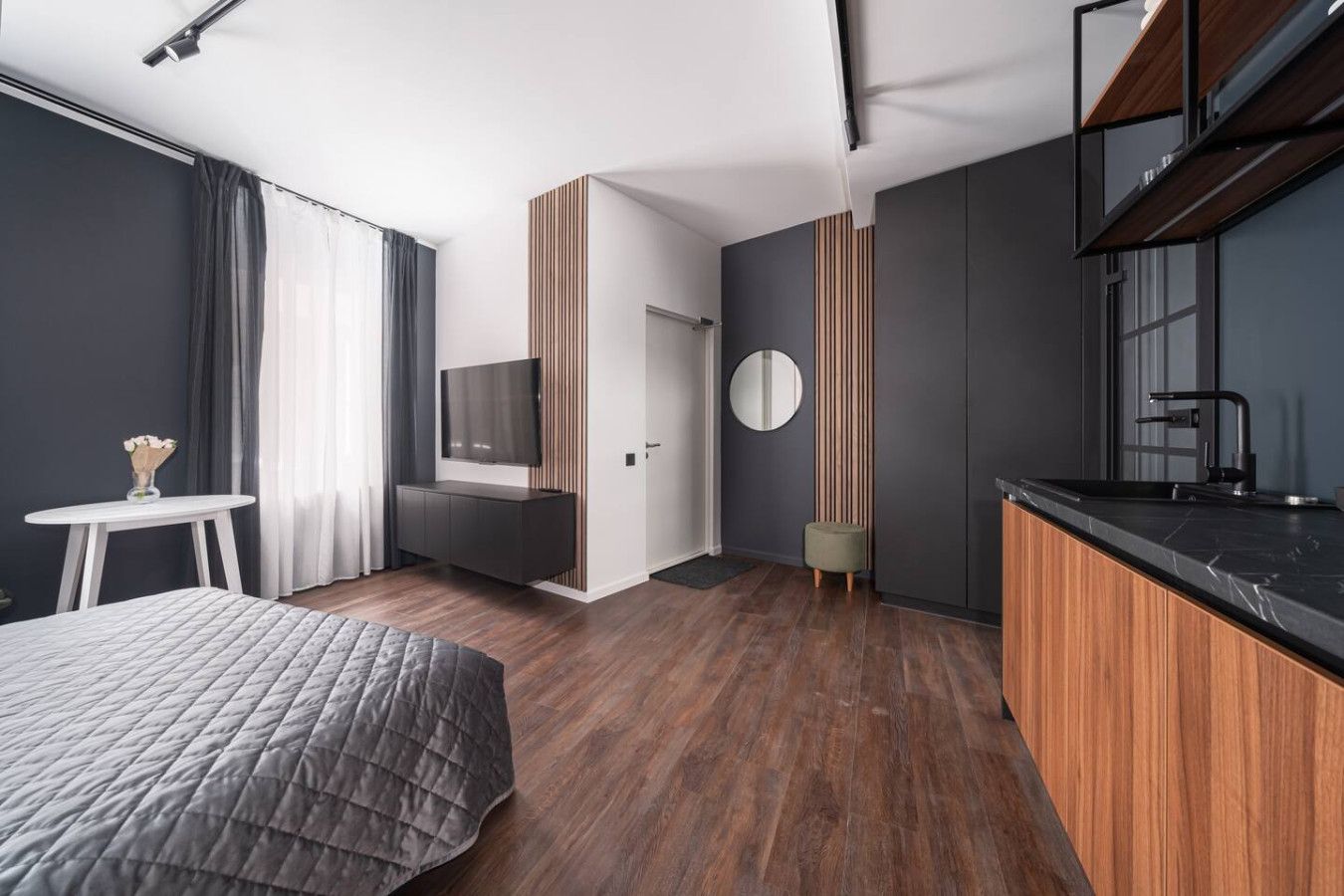 A modern hotel room with a bed, a small table with a vase, a TV, a round mirror, and a kitchenette.