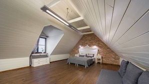 A bedroom with a sloped ceiling, wooden flooring, a bed with a white headboard, a gray sofa, and a small table with a lamp.