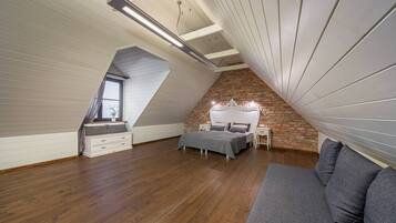 Ein Schlafzimmer mit schrägverdeckter Decke, Holzboden, einem Bett mit weißem Kopfteil, einem grauen Sofa und einem kleinen Tisch mit Lampe.