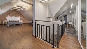 A spacious loft with a high ceiling, exposed brick walls, and a wooden floor. A modern bed and a dining area are visible.