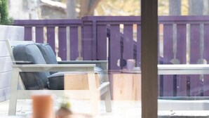 Un área de estar moderna al aire libre con una mesa y sillas de madera, barandal morado y vista a árboles y un edificio al fondo.