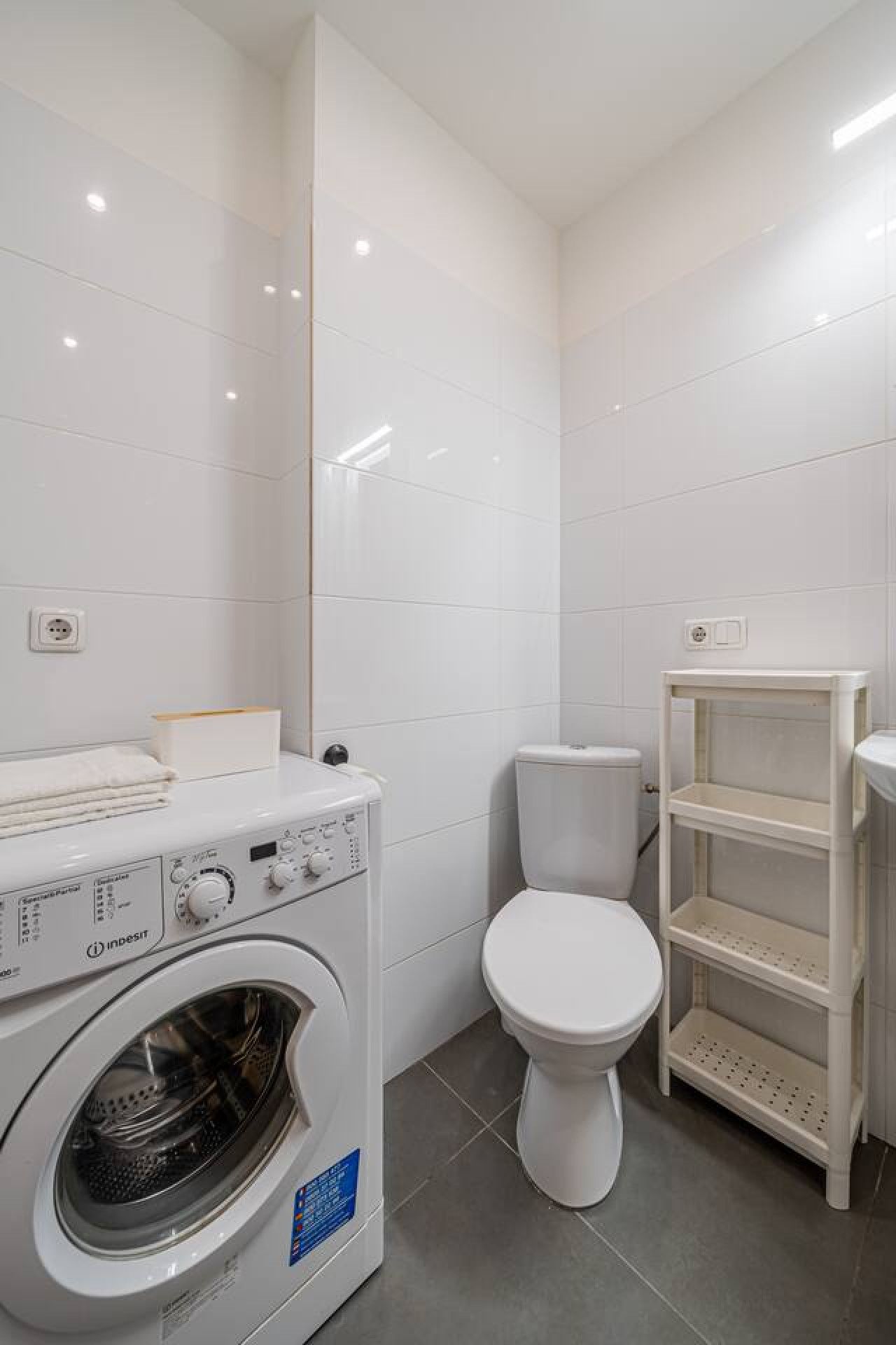 Une petite salle de bain équipée d’un lave-linge, de toilettes et d’une petite étagère.