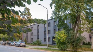 A modern building with a grey facade, multiple windows, and a parking lot with cars. - Trendy beach-front 1-bedroom apartment in Dzintari (Jūrmala)