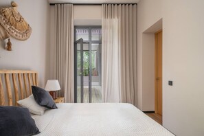 Un dormitorio con cama, cabecera de madera, mesita de noche con lámpara y vistas a un área exterior por medio de una puerta de vidrio.