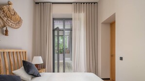 Quarto com cama, cabeceira de madeira, mesa de cabeceira com abajur e vista para área externa através de uma porta de vidro.