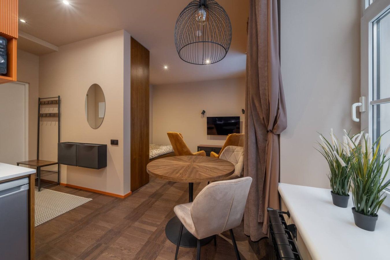 A compact hotel room with a round dining table, two chairs, a small sofa, a TV, and a potted plant.