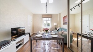 Un espace repas moderne avec une table en bois, des chaises transparentes et un téléviseur posé sur un meuble blanc.